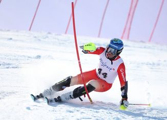 Ein Skifahrer, der einen blauen Helm, eine Brille und einen rot-weißen Rennanzug trägt, fährt durch ein Slalomtor, neigt sich stark und wirft Schnee auf einer sonnenbeschienenen Abfahrtsstrecke auf.
