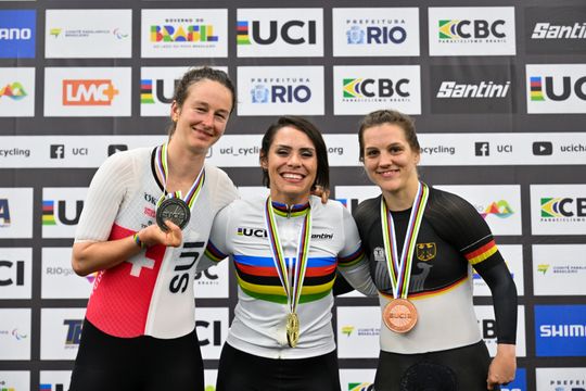 Drei Radsportlerinnen stehen lächelnd auf einem Podium, jede trägt eine Medaille. Die Athletin in der Mitte trägt ein Trikot mit Regenbogenstreifen und hält die Goldmedaille in der Hand, flankiert von den Silber- und Bronzemedaillengewinnern. Hinter ihnen sind die Logos der Sponsoren der Radsportveranstaltung zu sehen.