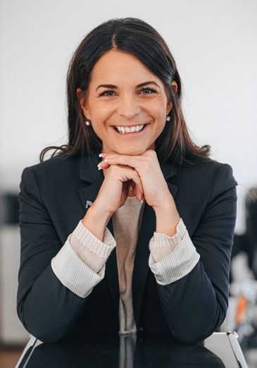 Eine Frau mit langen dunklen Haaren lächelt warm und stützt ihr Kinn auf ihre Hände. Sie trägt einen dunklen Blazer über einem hellen Pullover und sitzt in einem gut beleuchteten Raum.