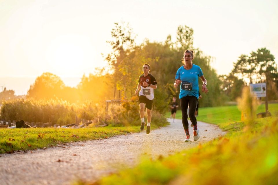 Zwei Läufer laufen bei Sonnenuntergang auf einem von Grün umgebenen Kiesweg um die Wette, mit Nummern an ihren Trikots. Die Sonne leuchtet hell im Hintergrund und wirft ein warmes Licht auf die Szene.