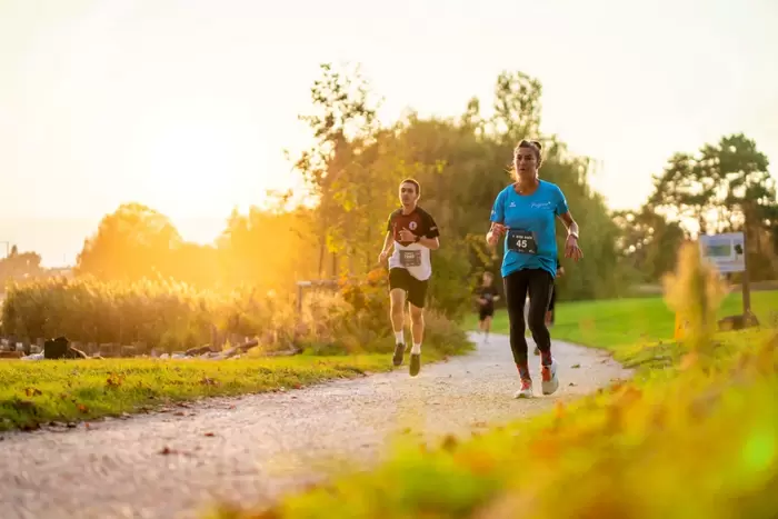 Zwei Läufer laufen bei Sonnenuntergang auf einem von Grün umgebenen Kiesweg um die Wette, mit Nummern an ihren Trikots. Die Sonne leuchtet hell im Hintergrund und wirft ein warmes Licht auf die Szene.