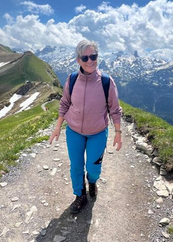 Eine Frau mit Sonnenbrille, rosa Jacke und blauer Hose wandert auf einem Bergpfad mit schneebedeckten Gipfeln und einem blauen Himmel mit Wolken im Hintergrund.