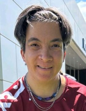 Eine Person mit kurzen braunen Haaren, die ein kastanienbraunes Sporttrikot und mehrere Halsketten trägt, lächelt im Freien in hellem Sonnenlicht mit einem modernen Gebäude im Hintergrund.