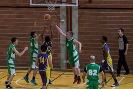 Eine Gruppe von Basketballspielern in grünen und lilafarbenen Trikots springt und greift nach dem Ball in der Nähe des Korbes auf einem Hallenplatz, während rechts ein Schiedsrichter zusieht.