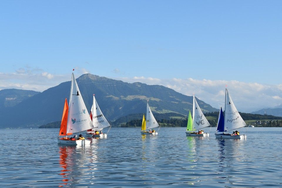 Sechs kleine Segelboote mit bunten Segeln gleiten auf einem ruhigen See mit Bergen und blauem Himmel im Hintergrund. Im Wasser spiegeln sich die Boote und die Landschaft, wodurch eine friedliche und malerische Szene entsteht.
