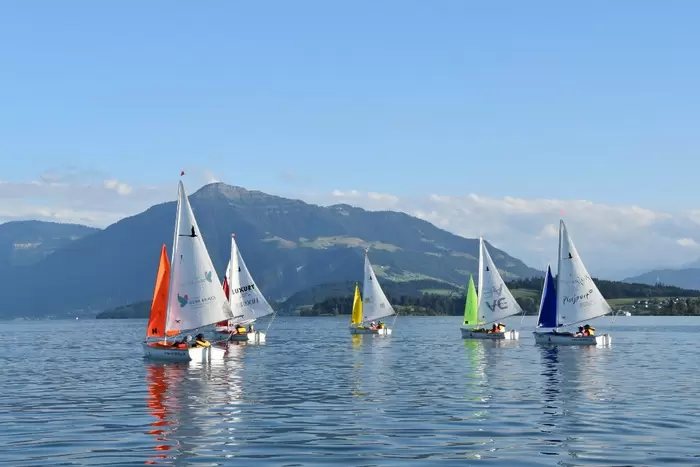 Sechs kleine Segelboote mit bunten Segeln gleiten auf einem ruhigen See mit Bergen und blauem Himmel im Hintergrund. Im Wasser spiegeln sich die Boote und die Landschaft, wodurch eine friedliche und malerische Szene entsteht.
