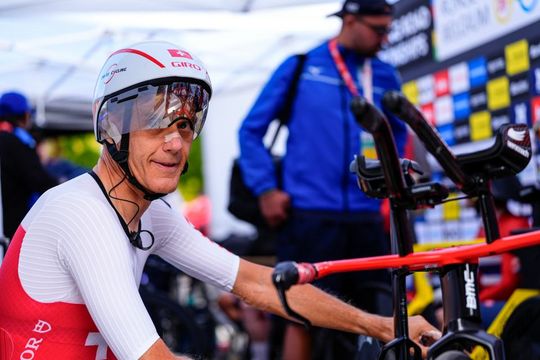 Ein Radfahrer in einem rot-weißen Anzug mit einem Visierhelm bereitet sich auf ein Rennen vor und sitzt auf einem Rennrad. Im Hintergrund sind andere Personen und eine Kulisse mit verschiedenen Logos zu sehen.