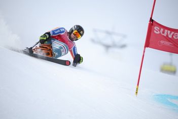 Ein alpiner Skifahrer mit Helm und Skibrille rast während eines Rennens einen verschneiten Hang hinunter, vorbei an einem Tor mit roter Flagge. Dahinter spritzt Schnee auf, und die Liftanlagen sind im Hintergrund verschwommen.