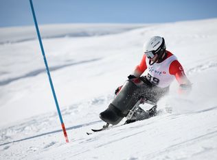 Ein Para-Alpin-Skifahrer mit Helm und Nummer 49 fährt mit einem Monoski einen verschneiten Hang hinunter und neigt sich stark, während der Schnee um ihn herum aufspritzt, wobei eine blaue Slalomstange sichtbar ist.