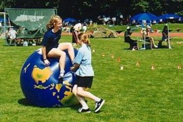 Zwei Mädchen spielen im Freien auf einer großen aufblasbaren Kugel auf einer Wiese, mit Menschen und Regenschirmen im Hintergrund, was auf eine Gemeinde- oder Schulveranstaltung hindeutet.