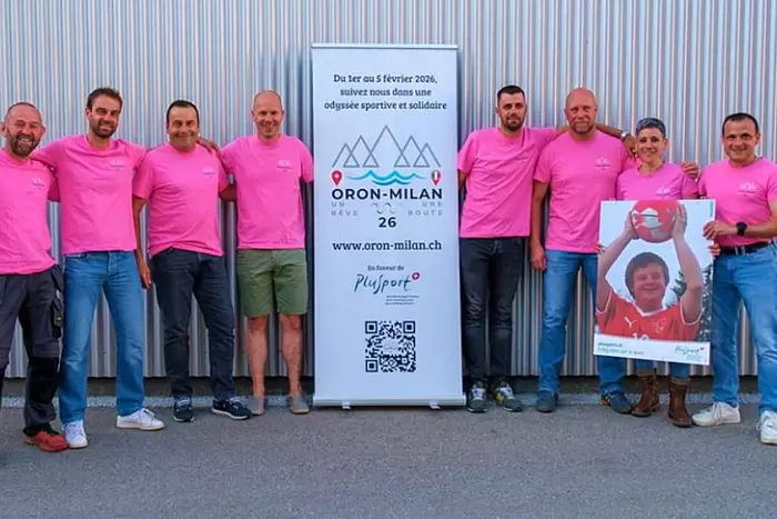 Acht Personen in passenden rosafarbenen Hemden stehen in einer Reihe im Freien und lächeln neben einem Banner und einer Frau, die ein großes Foto einer Person in einem roten Hemd hält, die einen Ball hält. Der Hintergrund ist eine Wand aus Wellblech.