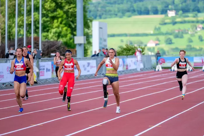 Fünf Sportlerinnen, darunter zwei mit Beinprothesen, sprinten während eines Rennens auf einer roten Bahn. Im Hintergrund sind an einem sonnigen Tag grüne Felder und Bäume zu sehen.
