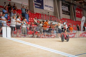 Ein Radfahrer mit Helm fährt auf einer Indoor-Radrennbahn, während die Zuschauer auf der Tribüne jubeln, klatschen und dem Fahrer hinter einer Glaswand die Hand reichen.