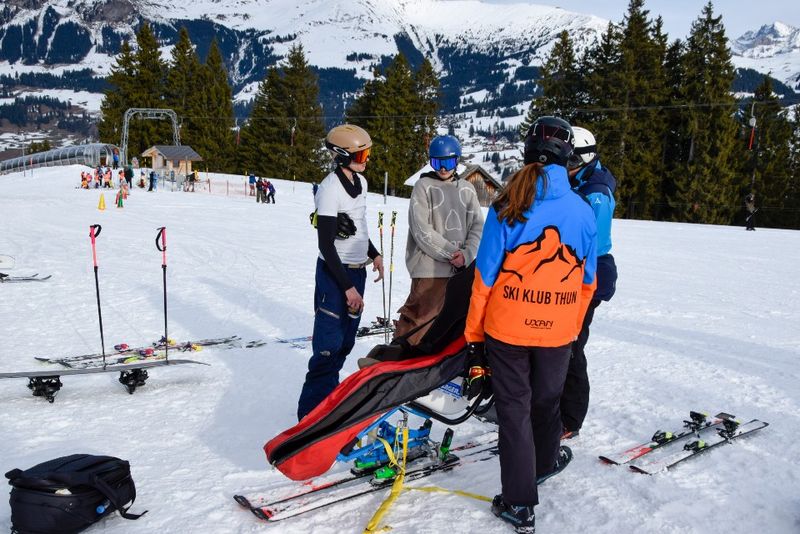 Eine Gruppe von Skifahrern, von denen einer eine orangefarbene Jacke des Skiclubs Thun trägt, steht um eine Person in einem Schlitten auf einem verschneiten Berghang, mit Skiern und Stöcken in der Nähe und Tannen im Hintergrund.