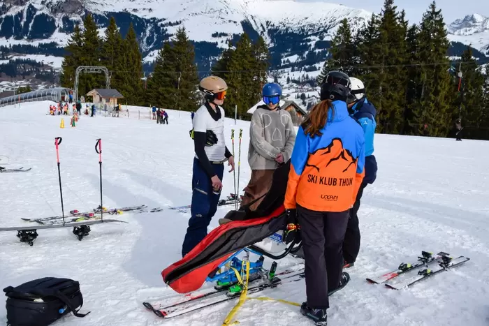 Eine Gruppe von Skifahrern, von denen einer eine orangefarbene Jacke des Skiclubs Thun trägt, steht um eine Person in einem Schlitten auf einem verschneiten Berghang, mit Skiern und Stöcken in der Nähe und Tannen im Hintergrund.