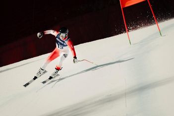 Ein Skifahrer in rot-weißem Anzug, Helm und Skibrille rast auf einer verschneiten Piste an einer roten Fahne vorbei bergab, wobei das Sonnenlicht Schatten auf den Schnee wirft.