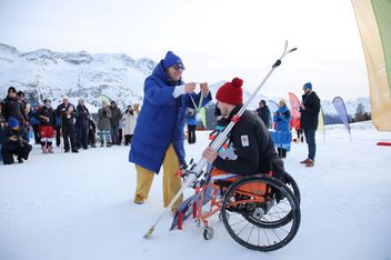 Eine Person im Rollstuhl, die Winterkleidung trägt, erhält eine Medaille von einer anderen warm gekleideten Person, umgeben von Schnee, Bergen und Zuschauern bei einer Outdoor-Veranstaltung.