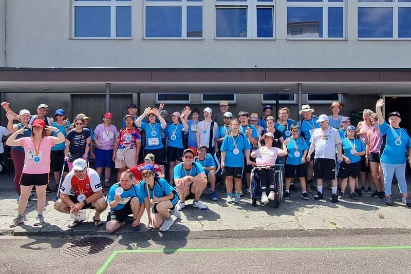 Eine große Gruppe von Menschen, viele mit Medaillen und blauen Hemden, posiert und winkt an einem sonnigen Tag vor einem Gebäude. Einige stehen, andere knien oder sitzen, lächeln und wirken glücklich nach einer Veranstaltung im Freien.