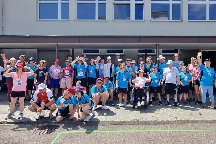 Eine große Gruppe von Menschen, viele mit Medaillen und blauen Hemden, posiert und winkt an einem sonnigen Tag vor einem Gebäude. Einige stehen, andere knien oder sitzen, lächeln und wirken glücklich nach einer Veranstaltung im Freien.