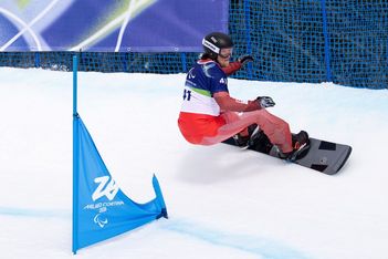 Ein Snowboarder mit Helm und Schutzbrille macht eine scharfe Kurve um eine blaue Fahne auf einer verschneiten Rennstrecke, wobei im Hintergrund ein Sicherheitsnetz und ein Banner zu sehen sind.
