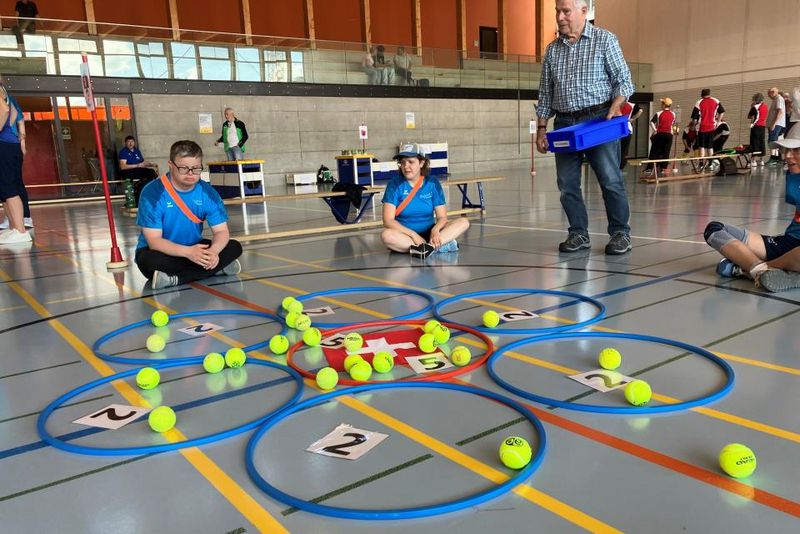 Menschen in blauen Hemden sitzen auf dem Boden einer Turnhalle um blumenförmig angeordnete Reifen, die mit Zahlen beschriftet sind. Tennisbälle sind in und um die Reifen herum verstreut. Ein älterer Mann steht in der Nähe und hält ein Tablett. Im Hintergrund sind weitere Personen zu sehen.