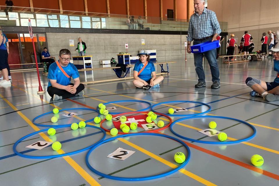 Menschen in blauen Hemden sitzen auf dem Boden einer Turnhalle um blumenförmig angeordnete Reifen, die mit Zahlen beschriftet sind. Tennisbälle sind in und um die Reifen herum verstreut. Ein älterer Mann steht in der Nähe und hält ein Tablett. Im Hintergrund sind weitere Personen zu sehen.