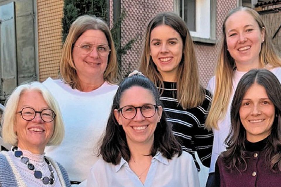 Sechs Frauen stehen im Freien eng beieinander und lächeln in die Kamera. Sie sind leger gekleidet und scheinen von unterschiedlichem Alter zu sein. Im Hintergrund ist ein Gebäude mit strukturierten Wänden zu sehen.