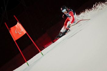 Ein Skifahrer in roter Ausrüstung und mit schwarzem Helm rast bei einem alpinen Skiwettkampf an einer roten Fahne mit dem paralympischen Symbol vorbei durch den Schnee.