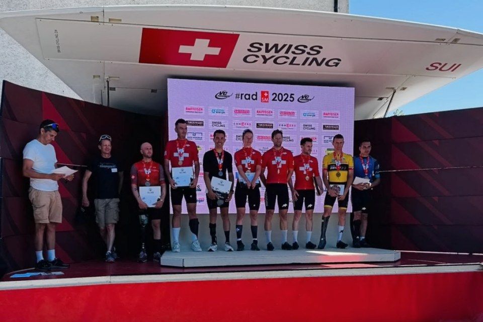 Sieben Radfahrer in roten Schweizer Trikots stehen auf einem Podium mit Medaillen und Urkunden an einer Swiss Cycling-Veranstaltung. Links davon stehen zwei Personen; im Hintergrund sind ein Swiss Cycling-Banner und Sponsorenlogos zu sehen.
