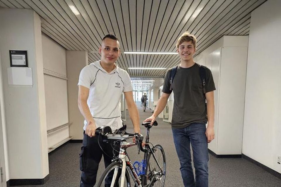 Zwei junge Männer stehen in einem Flur; einer hält ein Rennrad in der Hand. Beide lächeln, sind leger gekleidet und der Flur hat weiße Wände, einen dunklen Boden und eine gestreifte Decke.