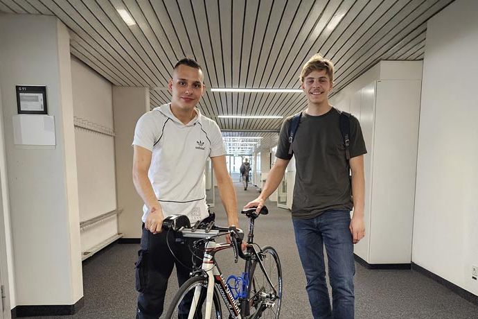Zwei junge Männer stehen in einem Flur; einer hält ein Rennrad in der Hand. Beide lächeln, sind leger gekleidet und der Flur hat weiße Wände, einen dunklen Boden und eine gestreifte Decke.