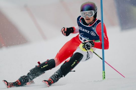 Ein Skifahrer in rot-weißer Ausrüstung macht während eines Rennens eine scharfe Kurve um eine Slalomstange auf einer verschneiten Piste. Er trägt einen Helm, eine Brille und eine nummerierte Startnummer.