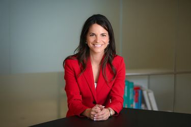 Eine Frau mit langen braunen Haaren, die einen roten Blazer und ein weißes Oberteil trägt, lächelt, während sie an einem schwarzen Tisch sitzt und die Hände verschränkt. Im Hintergrund sind bunte Bücher verschwommen zu sehen.