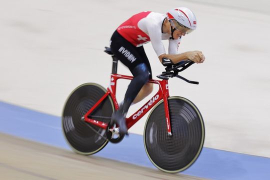 Ein Radfahrer in rot-weißer Uniform und mit Helm fährt auf einem roten Rennrad mit hoher Geschwindigkeit auf einer Indoor-Velodrom-Bahn. Der Hintergrund ist unscharf, um die Bewegung zu betonen.
