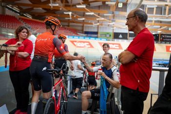 Ein Radfahrer in Orange schüttelt einem sitzenden Radfahrer in einer Radrennhalle die Hand, während andere in roten Hemden in der Nähe zuschauen. Die Umgebung wirkt energiegeladen und unterstützend.