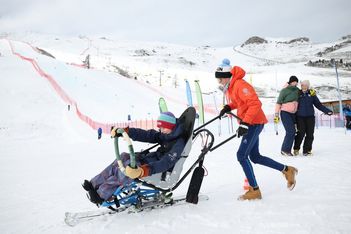 Eine Person auf einem Sitz-Ski wird von einem Skilehrer auf einer verschneiten Piste geschoben, während zwei Personen im Hintergrund zusehen. Skistöcke, Fahnen und schneebedeckte Berge sind unter einem bewölkten Himmel zu sehen.