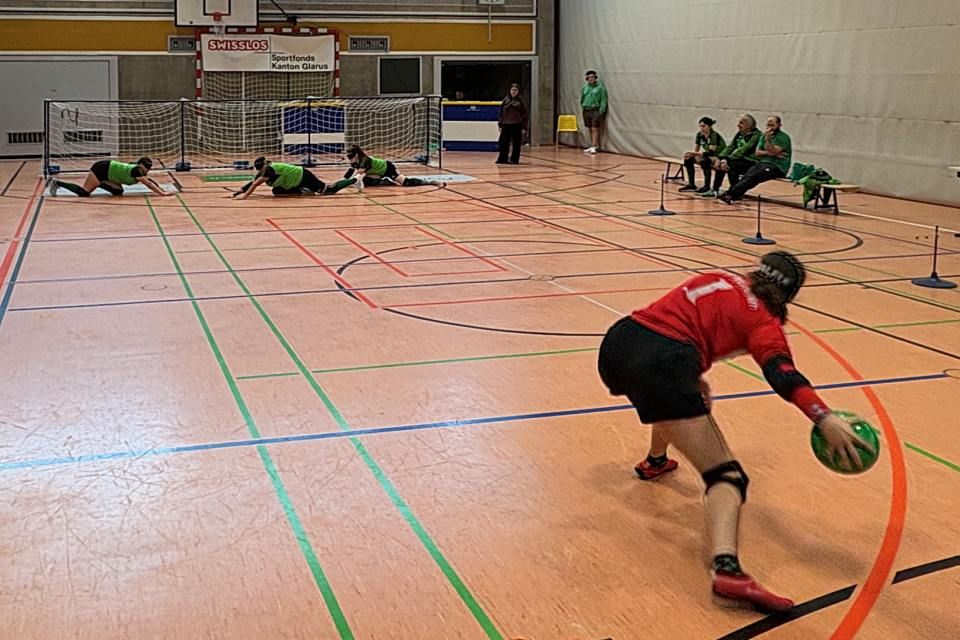 Ein Goalball-Spieler in einem roten Trikot bereitet sich auf den Wurf vor, während drei Spieler in grünen Trikots ein Tor auf einem Hallenplatz verteidigen. Zuschauer und Offizielle sitzen an der Seite.