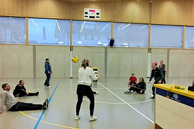 Eine Gruppe von Personen spielt Volleyball im Sitzen in einer Sporthalle. Die meisten Spieler sitzen auf dem Boden, während zwei in der Nähe des Netzes stehen und sich darauf vorbereiten, den Ball zu schlagen. Die Zuschauer schauen vom Rand aus zu.
