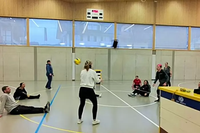 Eine Gruppe von Personen spielt Volleyball im Sitzen in einer Sporthalle. Die meisten Spieler sitzen auf dem Boden, während zwei in der Nähe des Netzes stehen und sich darauf vorbereiten, den Ball zu schlagen. Die Zuschauer schauen vom Rand aus zu.