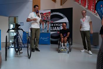 Drei Männer lächeln drinnen in der Nähe eines Handbikes und eines Rollstuhls; einer spricht in ein Mikrofon, einer sitzt in einem Rollstuhl und ein anderer steht in der Nähe. Im Hintergrund sind ein Banner und Fahrradplakate zu sehen.