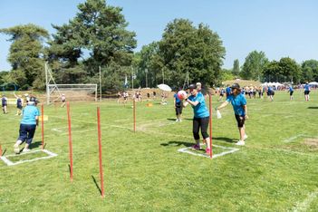 Menschen in blauen Hemden nehmen an einem Staffelspiel auf einem Sportplatz teil, bei dem sie sich einen Ball zuspielen, während andere im Hintergrund unter sonnigem Himmel zusehen oder spielen.