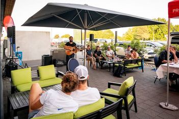 Ein Mann spielt Gitarre und singt unter einem großen Sonnenschirm auf einer Terrasse, während mehrere Personen auf Sofas und Stühlen sitzen, sich entspannen und zuhören. Im Hintergrund sind Autos und Bäume zu sehen.