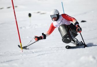 Ein Alpinskifahrer im rot-weißen Anzug fährt auf einem Monoski bergab, wobei er mit Hilfe von Auslegern durch ein Slalomtor auf einer verschneiten Piste navigiert.