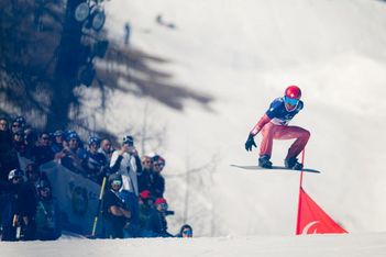 Ein Snowboarder mit rotem Helm und Brille befindet sich während eines Sprungs bei einem Schneewettbewerb in der Luft. In Winterkleidung gekleidete Zuschauer schauen vom Rand aus zu. Auf der Piste ist eine rote Flagge zu sehen.
