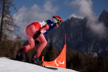Ein Skifahrer in rot-weißer Ausrüstung und mit Helm fährt auf einer verschneiten Piste bergab, vorbei an einer orangefarbenen Fahne mit Bergen und Bäumen im Hintergrund unter einem teilweise bewölkten Himmel.