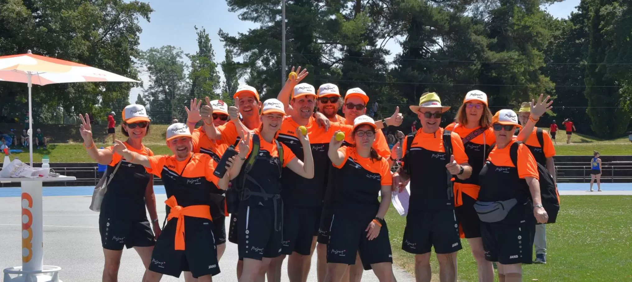 Eine Gruppe von Menschen in passenden orange-schwarzen Sportoutfits und Mützen posiert an einem sonnigen Tag im Freien, lächelt und winkt in die Kamera, während im Hintergrund Bäume und eine Sportbahn zu sehen sind.
