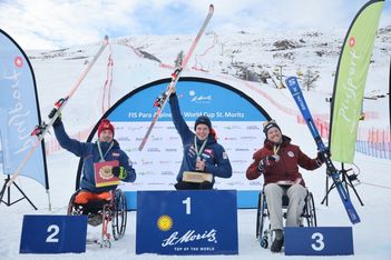 Drei männliche Athleten im Rollstuhl feiern auf einem verschneiten Podium beim FIS Para Alpine Ski World Cup in St. Moritz, mit Skiern und Medaillen in der Hand, mit verschneiten Bergen und Veranstaltungsbannern im Hintergrund.