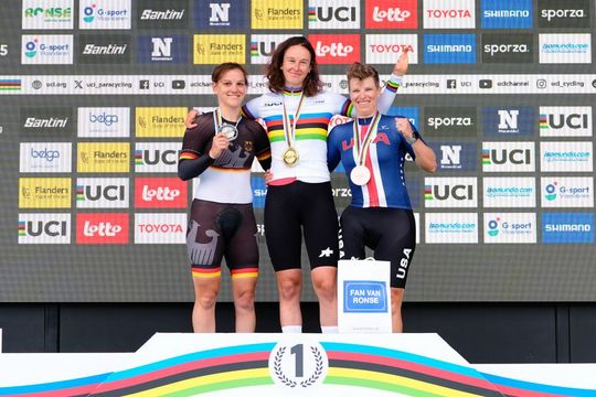 Drei Radfahrerinnen stehen lächelnd auf einem Podium und halten Medaillen in der Hand. Die mittlere Radfahrerin trägt ein Regenbogentrikot und eine Goldmedaille, flankiert von Silber- und Bronzemedaillengewinnern. Auf dem Hintergrund hinter ihnen sind Sponsorenlogos zu sehen.