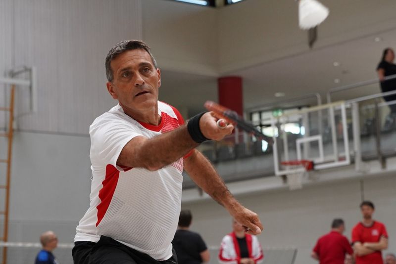 Ein Mann in einem weiß-roten Hemd spielt in einer Halle Badminton, wobei er seinen Schläger nach vorne streckt, um einen Federball zu schlagen. Im Hintergrund sind andere Spieler und ein Basketballkorb zu sehen.