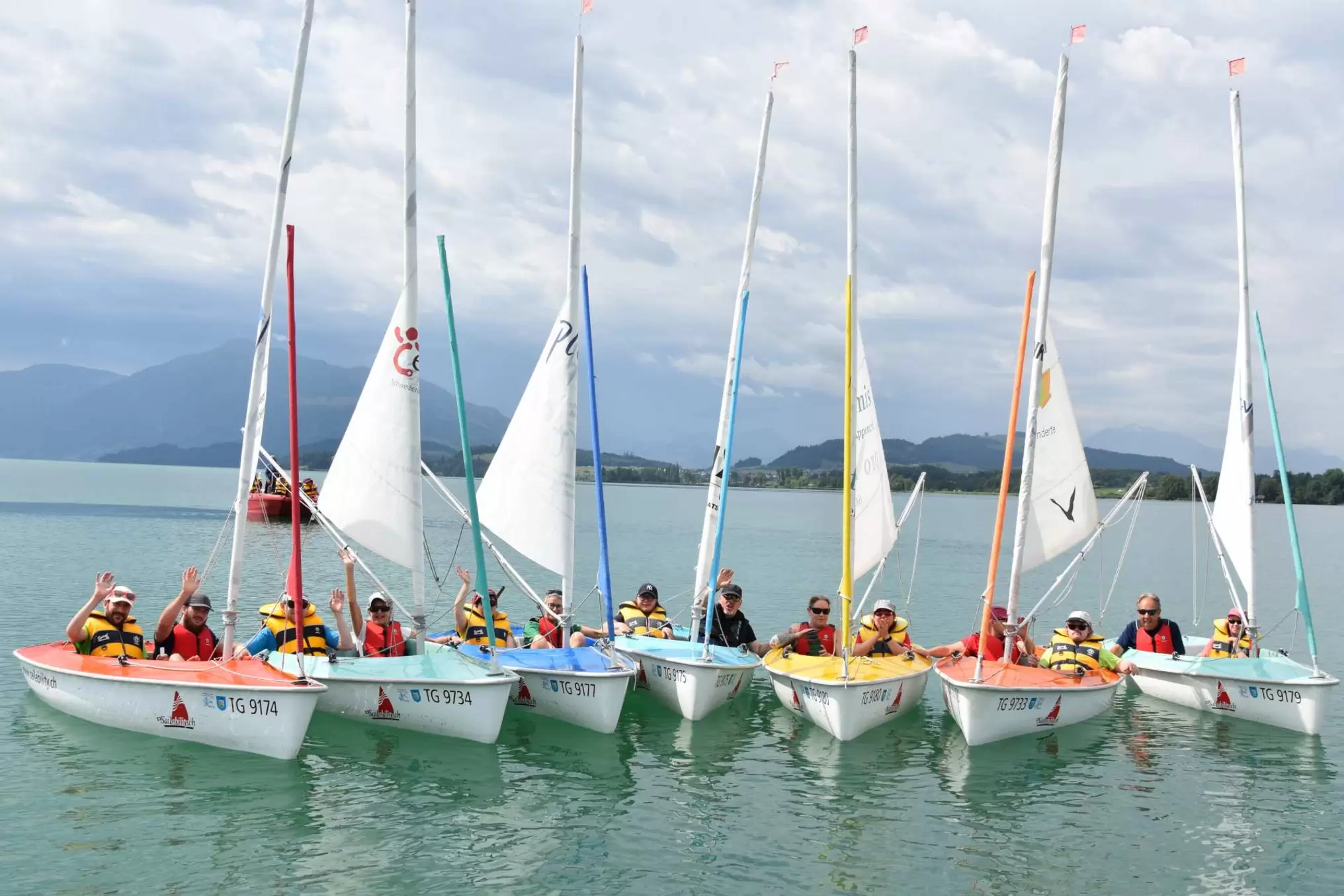 Eine Gruppe von Menschen mit Schwimmwesten sitzt in sechs kleinen Segelbooten auf einem ruhigen See, mit Bergen und wolkenverhangenem Himmel im Hintergrund.